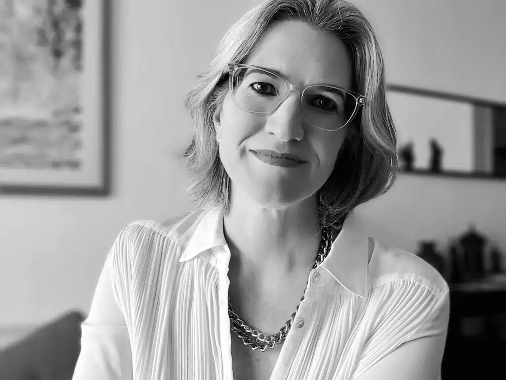 Foto de Suzeli Damaceno. Mulher branca, cabelos loiros acima dos ombros, olhos castanhos escuros. Usa óculos de armação grande e transparente, camisa branca e colares de metal.