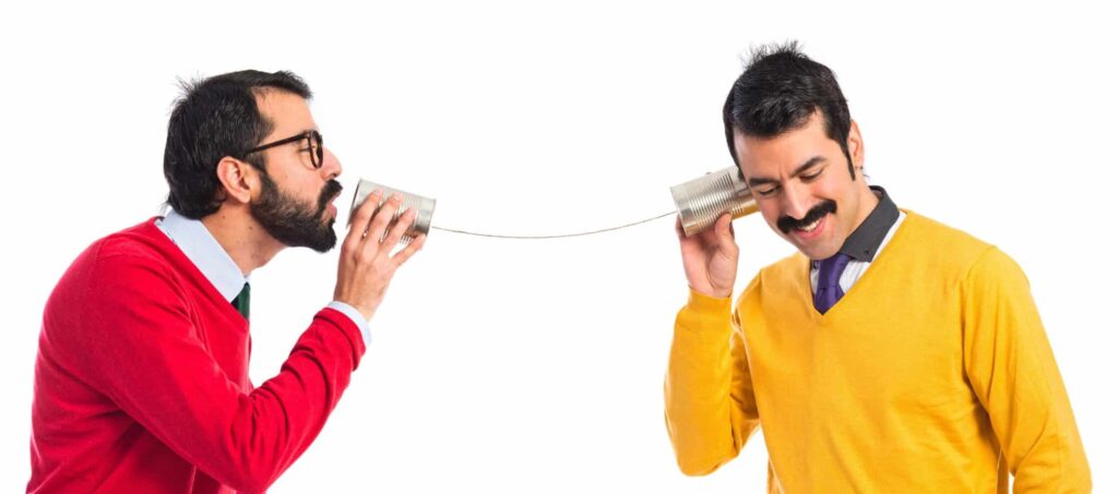 Foto de dois homens que simulam uma conversa utilizando duas latas parecidas com as de leite condensado que estão unidas por um barbante. Elas simulam um telefone.