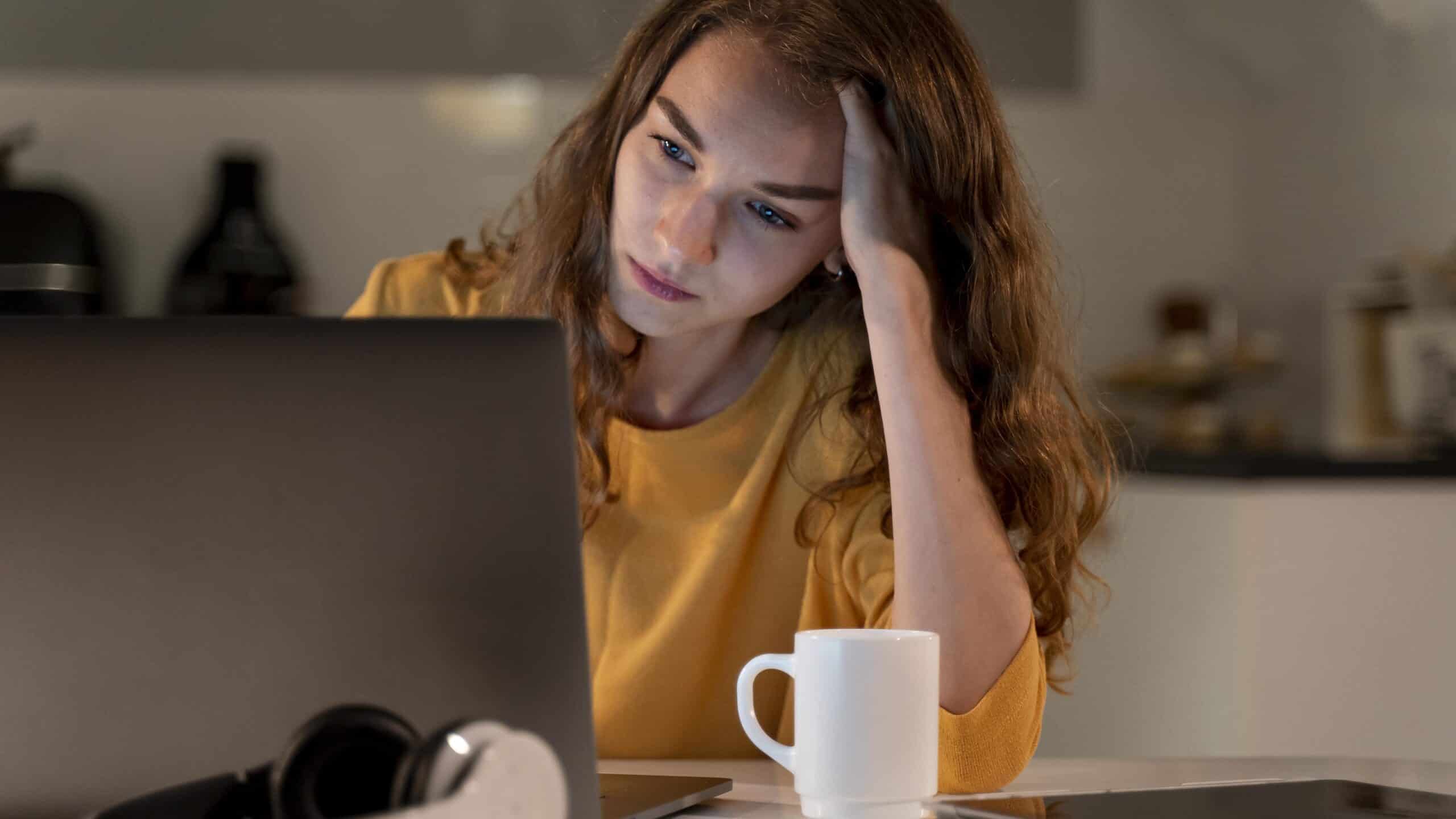 Foto de uma mulher em frente a um laptop, que está sob uma mesa com alguns objetos. Ela apoia levemente sua cabeça em uma das mãos e olha para a tela do computador com expressão séria e desanimada.