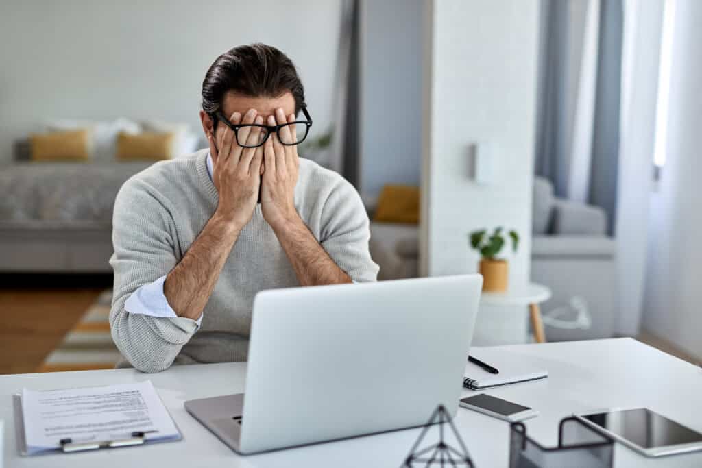 Foto de homem sentado à frente de uma mesa com laptop aberto, celular, tablet e alguns papéis. Ele está com os cotovelos apoiados na mesa e com as mãos cobrindo o rosto por debaixo dos óculos. O rapaz veste roupa formal e está em um ambiente que parece um quarto.