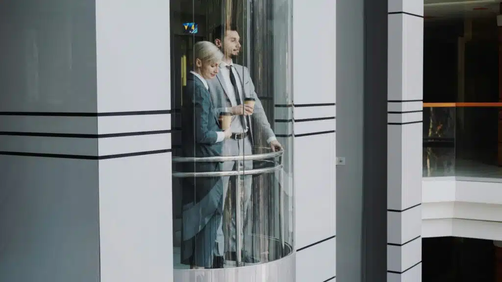 Um homem e uma mulher com trajes formais num elevador panorâmico em prédio corporativo. Estão lado a lado olhando para além do vidro.