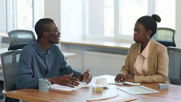 Duas pessoas negras em trajes formais conversam ao redor de uma mesa com papéis, xícaras e outros objetos de escritório.